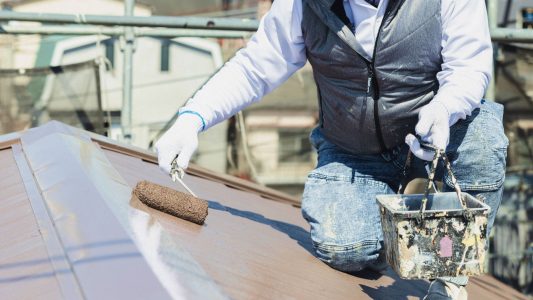 屋根の上で塗装作業を行う職人。ローラーを使って金属製の屋根を茶色系の塗料で丁寧に塗っている様子。