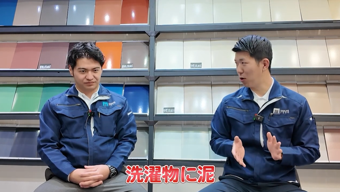 外壁塗装の専門家2名が、高圧洗浄時に発生した「洗濯物に泥」が付着するというクレーム事例について解説している動画のキャプチャ。洗浄水が近隣に飛散するリスクと、事前の声かけや養生の重要性を説明しているシーン。
