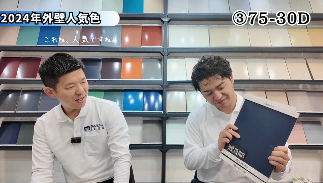 外壁塗装の専門家2名が、2024年の外壁人気色ランキング第3位「75-30D」について解説している動画の一場面。右の男性が落ち着いた濃い青色のカラーサンプルを持ち、左の男性が「これね、人気ですね」とコメントしている。背景には豊富な色見本が並び、プロが最新のトレンドを伝える、信頼感のある色選びのコンサルティングシーン。