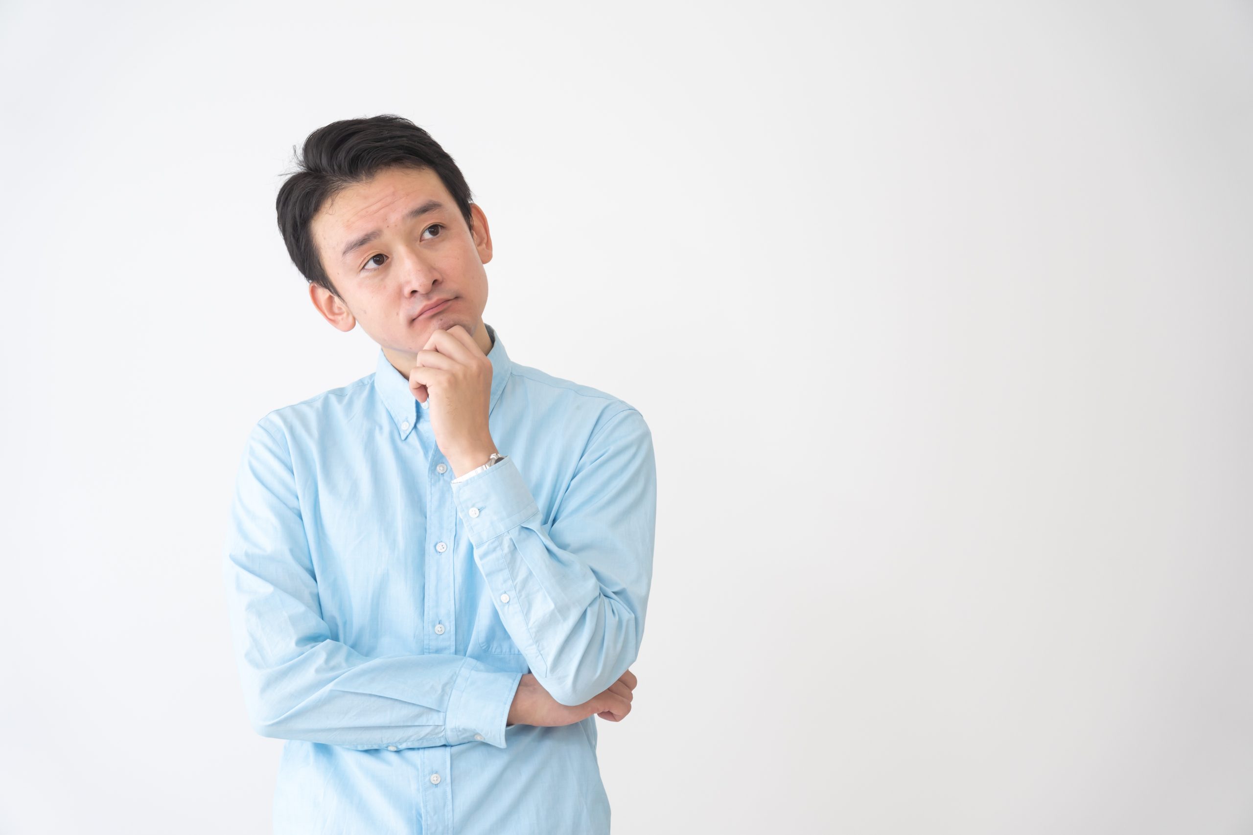 水色のシャツを着た若い男性が、白い背景をバックに、腕を組んで顎に手を当て、難しい顔で考え込んでいる。外壁塗装における1液塗料と2液塗料の違いや、価格、耐久性などのメリット・デメリットを比較検討し、どちらを選ぶべきか悩んでいる施主を表現。後悔しない塗料選びの疑問や不安を象徴する、共感性の高いイメージ。