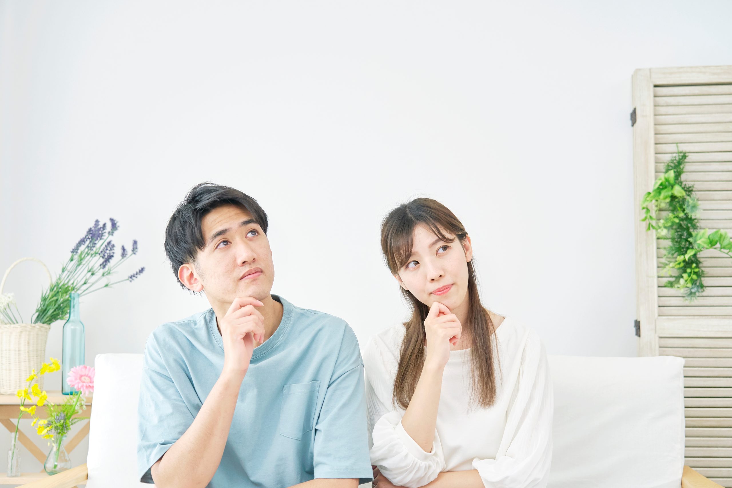 明るいリビングのソファに座り、若い夫婦が揃って顎に手をあてて考え込んでいる。外壁塗装の塗料選びや色選びで後悔しないために、1液塗料と2液塗料のどちらが良いかなど、性能や価格を比較検討している様子を象徴する一枚。マイホームの将来について真剣に悩む、共感性の高いイメージ。