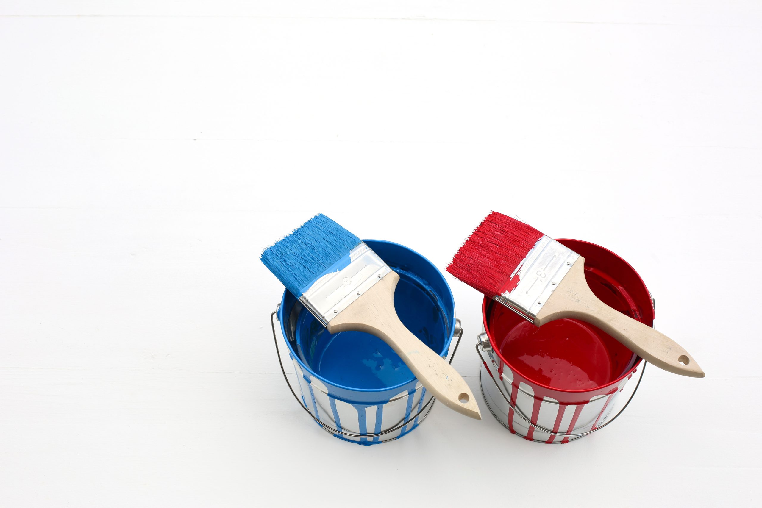 白い背景に並べられた、赤と青の2つのペンキ缶とハケ。外壁塗装における1液塗料と2液塗料の違いを比較するコンセプトを象徴する一枚。それぞれの塗料が持つ性能、耐久性、価格などのメリット・デメリットを検討し、最適な選択をすることの重要性を示す、クリーンで分かりやすいイメージ。
