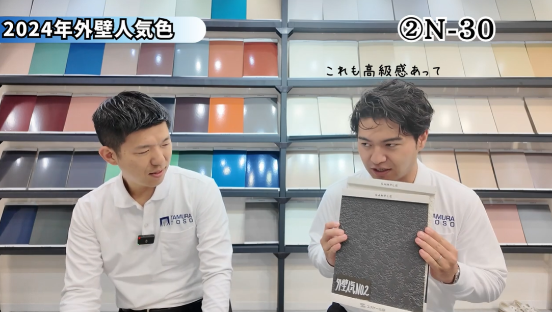 外壁塗装の専門家2名が、2024年の外壁人気色ランキング第2位「N-30」を紹介しているYouTube動画のキャプチャ。右の男性が、表面に凹凸のある黒に近いダークグレーのサンプルボードを手に持ち、「これも高級感あって」と色の持つ魅力を説明している。外壁の色選びで重要な、色の持つ雰囲気や質感をプロが解説する場面。